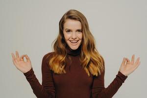 Confident woman in a brown turtleneck making OK gestures with both hands, her joyful demeanor against a neutral background photo