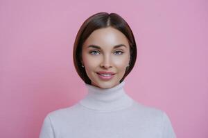 Elegant woman with glossy hair in a turtleneck sweater, showcasing a subtle smile, against a pink backdrop photo