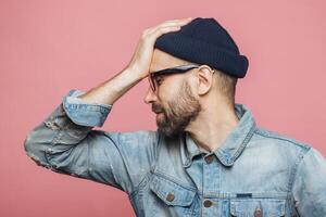 Man in denim jacket, wiping forehead with hand in a relieved gesture, wearing beanie and glasses, against a pink background photo