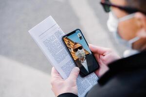 hombre en un vídeo llamada durante COVID-19, exhibiendo pandemia vida con un teléfono en mano, papel, mascarilla, y reflexión en lentes foto