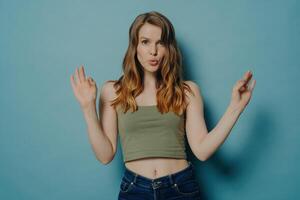Woman in green top making a playful face and OK sign with both hands, standing against a calm blue background photo