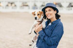 Cheerful woman in blue attire holding a playful Jack Russell Terrier on a sandy beach, both looking ahead with bright, happy eyes photo