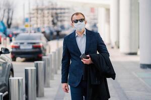 Professional man in a blue checkered suit walks confidently in the city, wearing a protective face mask and sunglasses, holding a phone. photo