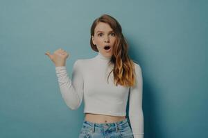 Shocked young woman with a hand gesture pointing sideways, expressing surprise and astonishment on a cool blue background photo