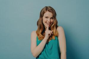 Playful young woman in a teal tank top making a shushing gesture, her eyes sparkling with fun against a blue backdrop photo