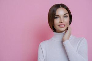 Radiant young lady with lustrous hair in a white turtleneck, hand on cheek, against a vibrant pink background photo