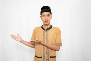 Asian Muslim man wearing brown Muslim clothes smiling happily while pointing at an empty space. Isolated white background. photo