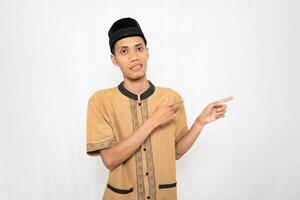 Asian Muslim man wearing brown Muslim clothes smiling happily while pointing at an empty space. Isolated white background. photo