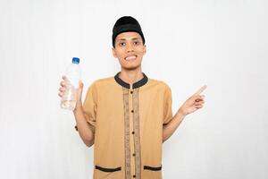 Asian Muslim man wearing Muslim clothes carrying mineral water in a bottle while pointing to the empty space. Isolated white background. photo