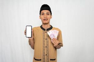 Asian Muslim man wearing brown Muslim shirt smiling happily while holding money and showing smartphone screen. Isolated white background. photo