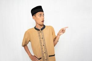 Asian Muslim man wearing brown Muslim clothes smiling happily while pointing at an empty space. Isolated white background. photo