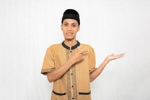 Asian Muslim man wearing brown Muslim clothes smiling happily while pointing at an empty space. Isolated white background. photo