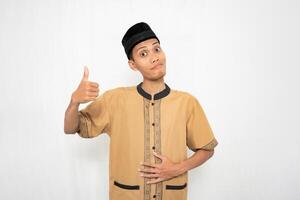 Asian Muslim man wearing Muslim clothes smiling happily, thumbs up gesture while holding a healthy or full stomach. Isolated white background. photo