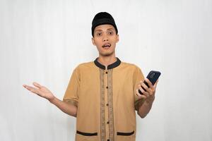 Asian Muslim man wearing Muslim clothes is shocked, confused and surprised while carrying a smartphone. Isolated white background. photo