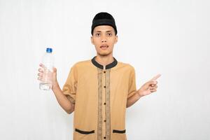 Asian Muslim man wearing Muslim clothes carrying mineral water in a bottle while pointing to the empty space. Isolated white background. photo