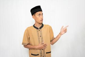 Asian Muslim man wearing brown Muslim clothes smiling happily while pointing at an empty space. Isolated white background. photo