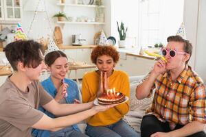 Make a wish. Woman wearing party cap blowing out burning candles on birthday cake. Happy Birthday party. Group of friends wishes girl happy birthday. People celebrating birthday with party at home photo