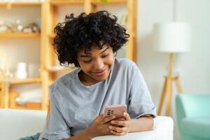 African girl holding smartphone touch screen typing scroll page at home. Woman with cell phone surfing internet using social media apps playing game. Shopping online Internet news cellphone addiction photo