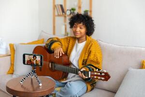 Blogger guitarist. African american girl blogger playing guitar talking to webcam recording vlog. Social media influencer woman streaming at home indoors. Music content creator broadcast tutorial photo