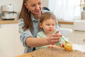 contento familia a hogar. madre alimentación su bebé niña desde cuchara en cocina. pequeño niñito niño con sucio gracioso cara come sano comida a hogar. joven mujer mamá dando comida a niño hija foto