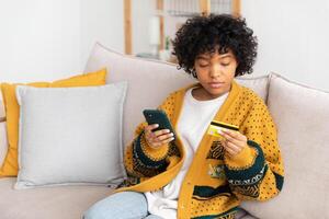 africano americano mujer compras en línea participación teléfono inteligente pago con oro crédito tarjeta. niña sentado a hogar comprando en Internet entrar crédito tarjeta detalles. en línea compras comercio electrónico entrega Servicio foto