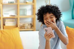 Excited happy young black african american woman holding cell phone laughing feeling joy getting mobile message. Overjoyed girl laughing aloud sitting on couch watching funny video reading news photo