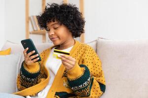 africano americano mujer compras en línea participación teléfono inteligente pago con oro crédito tarjeta. niña sentado a hogar comprando en Internet entrar crédito tarjeta detalles. en línea compras comercio electrónico entrega Servicio foto