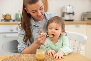 contento familia a hogar. madre alimentación su bebé niña desde cuchara en cocina. pequeño niñito niño con sucio gracioso cara come sano comida a hogar. joven mujer mamá dando comida a niño hija foto