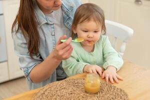 contento familia a hogar. madre alimentación su bebé niña desde cuchara en cocina. pequeño niñito niño con sucio gracioso cara come sano comida a hogar. joven mujer mamá dando comida a niño hija foto