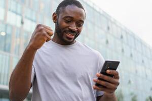 contento africano americano hombre eufórico ganador con teléfono inteligente en calle en ciudad. persona chico mirando a célula teléfono leyendo genial Noticias consiguiendo bueno resultado victorioso en línea oferta sensación asombrado. victorioso gesto foto