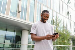 African american man holding smartphone touch screen typing scroll page on urban street in city. Guy with cell phone surfing internet using social media apps shopping online. Cellphone addiction photo