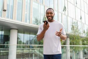 African american man talking on smartphone on street in city outdoor. Man with cell phone chatting with friends. Smiling person making answering call by cellphone. Guy having conversation by mobile photo