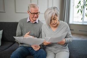 Sad tired disappointed middle aged senior couple sit with paper document. Unhappy older mature man woman reading paper bill managing bank finances calculating taxes planning loan debt pension payment photo