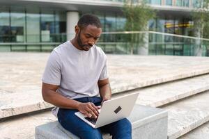 africano americano hombre persona de libre dedicación utilizando ordenador portátil mecanografía chateando en urbano calle en ciudad. chico teniendo virtual reunión en línea charla conferencia. empresario programador trabajando en ordenador portátil al aire libre. remoto trabajo foto