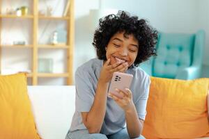 Excited happy young black african american woman holding cell phone laughing feeling joy getting mobile message. Overjoyed girl laughing aloud sitting on couch watching funny video reading news photo