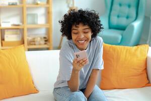 African girl holding smartphone touch screen typing scroll page at home. Woman with cell phone surfing internet using social media apps playing game. Shopping online Internet news cellphone addiction photo