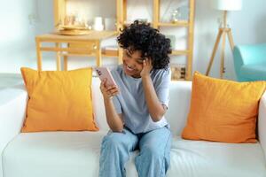 Excited happy young black african american woman holding cell phone laughing feeling joy getting mobile message. Overjoyed girl laughing aloud sitting on couch watching funny video reading news photo