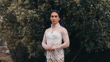 Elegant person in a lace dress standing before lush greenery, exuding a serene and sophisticated aura. video