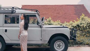 Elegant woman posing beside a classic off-road vehicle with a rustic house and greenery video