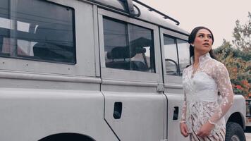 Elegant woman in a lace dress standing beside a vintage van, looking away thoughtfully. video