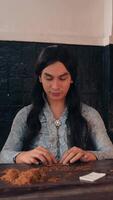 Serious young adult with long hair sitting at a table with a rustic backdrop looking at the camera with a neutral expression., wearing a patterned shirt video