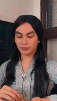Serene young adult with long hair, wearing a patterned shirt and pendant, focused on a handcraft video