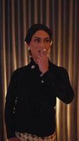 Elegant person in black attire with dramatic makeup posing with a cigarette against a curtained backdrop. video