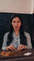 Serious young adult with long hair sitting at a table with a rustic backdrop looking at the camera with a neutral expression., wearing a patterned shirt video