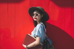 ai generado asiático mujer participación un maleta con un libro en contra rojo pared foto