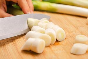 Woman's hand holding a knife cutting fresh Japanese scallions on a wooden chopping board. Sliced leek or Chopped green japanese bunching onion on a wooden board. photo