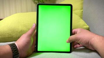 A man's hand holds a digital tablet with a blank green screen. A man is relaxing at home and playing computer video games and browsing a website.