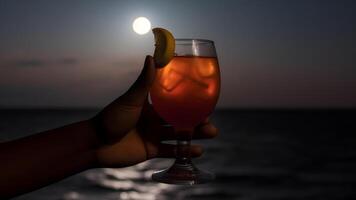 ai generado africano americano mano participación vaso de cóctel en borroso mar horizonte antecedentes a lleno Luna noche, neural red generado imagen foto