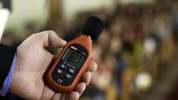 Close up of a man hand holding a red sound level meter and analyzer on clapping audience background. Stock footage. Man measuring the noise level on blurred crowd background. photo