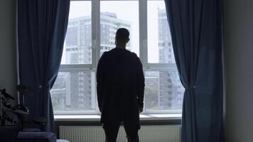 Thoughtful man coming to a window at home and looking through glass. Media. Man waiting for something at home in a room with blue curtains. photo